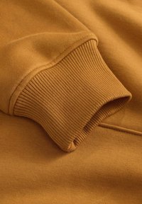 Tejido de sudadera de algodón en un cálido color mostaza, con puños acanalados y una textura suave. Detalles de costura visibles a lo largo de la manga.