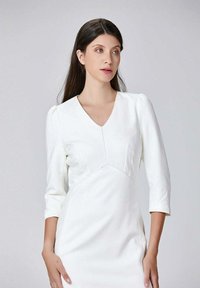 Robe blanche avec des manches trois-quarts, décolleté en V, corsage ajusté et détails de coutures. Tissu lisse au design minimaliste.