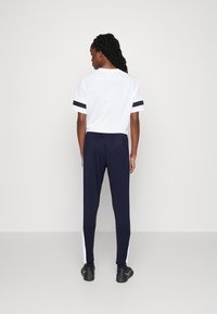 Camicia bianca a maniche corte con dettagli a strisce nere abbinata a pantaloni fitted blu scuro con strisce laterali bianche. Scarpe da ginnastica nere.