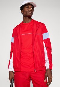 Κόκκινο αθλητικό σακάκι με μπλε και λευκές λεπτομέρειες, διαθέτει φερμουάρ και αντίστοιχο κόκκινο πουκάμισο από κάτω. Ελαφρύ, λείο ύφασμα.