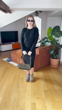 Vestido preto de mangas longas, até ao joelho, com silhueta ajustada. Botas cinzentas até ao tornozelo. A segurar uma pequena bolsa preta. Piso de madeira e fundo com planta verde.