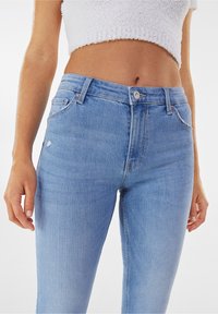 Jeans de mezclilla de color azul claro con una cintura alta, cinco bolsillos, cierre de cremallera con botón y detalles sutilmente desgastados en las piernas.