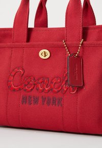 Červená plátenná taška s výšivkou textu "Coach" a "New York" v kontrastných farbách, zlaté kovanie a detail s visačkou.