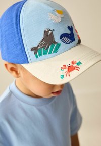 Blå og cremefarvet kasket med broderede havdyrsdesigns, herunder en sæl, hval og krabbe. Har en struktureret skygge og tekstiloverflade.