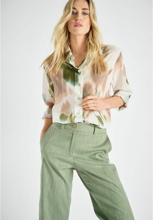 Blondine draagt een loszittend, knoopjes multicolor tie-dye shirt met opgerolde mouwen en een lichtgroene broek met hoge taille, poseert tegen een witte achtergrond.