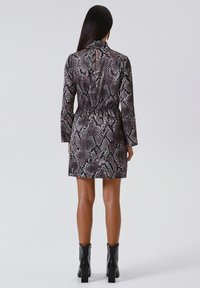 Kleid mit kurzen Ärmeln im Schlangenprint-Muster, mit hohem Ausschnitt, Taillenbindung und einer Schlüsselaugenöffnung hinten. Kombiniert mit schwarzen Knöchelstiefeln.