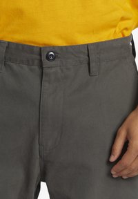 Pantaloni gri din bumbac cu închidere prin buton și față plată, având o textură netedă și buzunare laterale. Mână vizibilă pe talie.