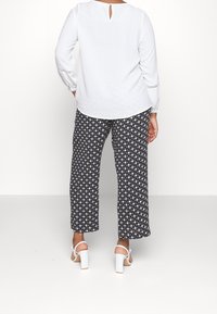 Persoon in een witte blouse met lange mouwen, zwart-wit gestreepte broek en witte hakken, staand met handen in de zakken.
