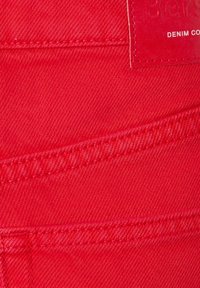 Tela de mezclilla roja con una textura suave, con costuras blancas contrastantes y una etiqueta de cuero con la inscripción "DENIM CO." para la identificación de la marca.