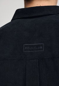 Czarna koszula z kordurowego materiału o teksturowej powierzchni i kołnierzyku, z naszywką z napisem "POLO CLUB" z tyłu.