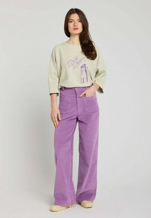 Femme debout portant un haut beige oversize avec un imprimé "Dirty Dancing" et un pantalon à jambes larges violet en velours côtelé taille haute.
