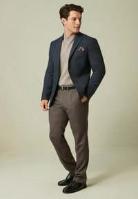 Blazer blu navy, maglione grigio chiaro e pantaloni marroni su misura completano i mocassini neri. Un fazzoletto da taschino notevole aggiunge un tocco di colore.