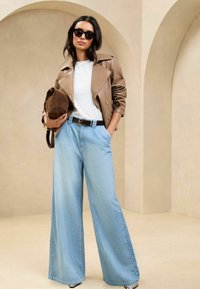 Chaqueta de cuero marrón con cinturón, camiseta blanca, vaqueros de pierna ancha azul claro, gafas de sol negras en forma de ojo de gato y bolso de ante marrón.