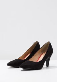 Zapatos de tacón alto de ante negro con punta afilada y un tacón moderado. El interior cuenta con un forro tostado con un acento rojo.