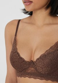 Bralette de encaje marrón con patrón floral, material semi-transparente, tirantes ajustables y diseño con borde en forma de ondas.