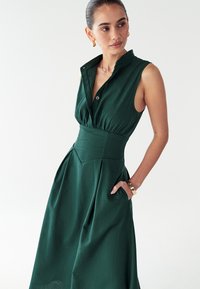 Grünes, ärmelloses Kleid mit kragenloser Knopfleiste, tailliertem Schnitt und gefälteltem Rock. Hergestellt aus strukturiertem Stoff, mit seitlichen Taschen und V-Ausschnitt.