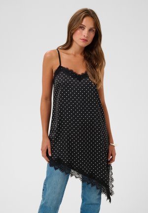 Jeune femme aux longs cheveux bruns porte une robe noire à pois avec bordure en dentelle par-dessus un jean bleu, debout devant un fond uni.