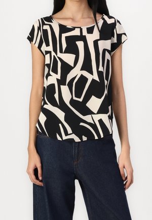 Kvinde iført en sort og beige bluse med abstrakt mønster og korte ærmer, kombineret med mørkeblå denimbukser, stående mod en ensfarvet baggrund.