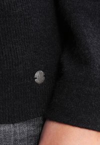 Maglione nero a maglia con un tessuto texturizzato. Presenta un bottone tondo in argento con un pattern a grana di legno. Aspetto liscio e morbido.