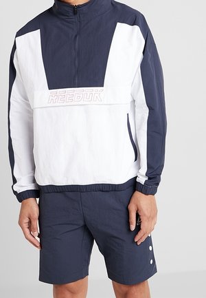 Man draagt een navyblauwe en witte Reebok windbreaker met halve rits en bijpassende navyblauwe shorts met knoopdetails aan de zijkant.