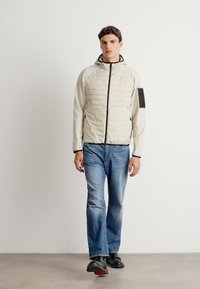 Light grey puffer jacket with a textured front, black accents, and a hood; paired with straight-leg blue jeans and black shoes.
