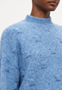 Suéter de punto azul claro con un alto cuello, que presenta un patrón texturizado y un acabado suave y esponjoso. El detalle enfatiza la tela acogedora y el diseño.
