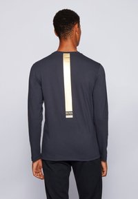 Dunkelblaues Langarm-T-Shirt mit einem vertikalen goldenen Streifen auf der Rückseite, einem Logo am unteren Ende und einer glatten, leichten Textur.