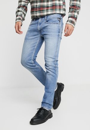 Mężczyzna ubrany w jasnoniebieskie obcisłe jeansy, czarne sznurowane buty oraz koszulę w kratę z podwiniętymi rękawami, stojący na jednolitym tle.