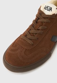 Smeđa tenisica od brušene kože s zaobljenim vrhom, smeđim vezicama, smeđim gumenim potplatom i vidljivim vunenim postavom s logotipom "VEJA" na jeziku.