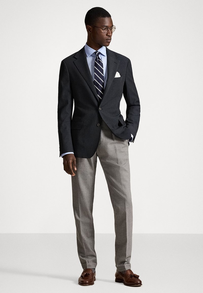 Σκούρο προσαρμοσμένο σακάκι, ανοιχτό πουκάμισο, ριγέ γραβάτα, γκρίζες παν trousers. Αξεσουάρ περιλαμβάνουν μαντήλι τσέπης και στρογγυλά γυαλιά. Καφέ loafers.