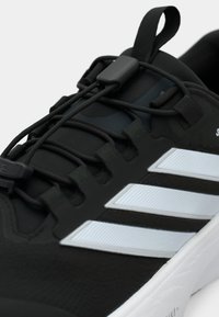 Μαύρο αθλητικό παπούτσι με ελαστικά κορδόνια, λαστιχένια σόλα και τρεις λευκές ρίγες στο πλάι· υλικό με υφή στο επάνω μέρος.