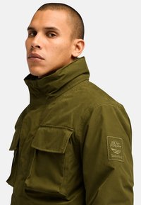 Chaqueta impermeable de color verde oliva con un alto cuello, dos bolsillos de solapa y un logo bordado en el hombro. Textura de tela duradera.