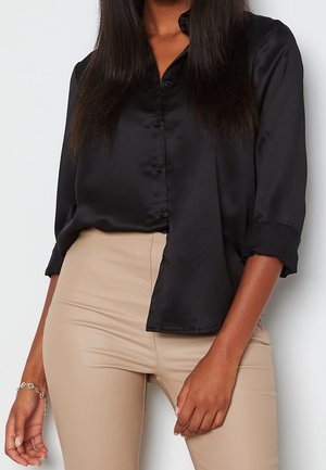 Femme portant un chemisier en satin noir à manches longues et un pantalon beige taille haute, une main reposant près de sa hanche.