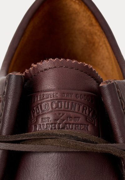 Mocasines de cuero marrón con una superficie texturizada, detalles cosidos y una etiqueta de marca. El interior es de ante en un tono beige.