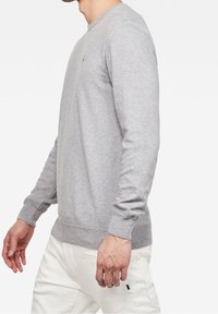 Harmaa neulepaita, jossa on pyöreä kaula-aukko, ribattu hihansuu ja helma. Paidassa on pieni logo rinnassa, ja se on valmistettu pehmeästä, teksturoidusta kankaasta.