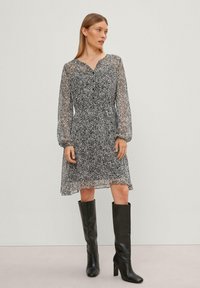 Zwart-witte bloemenprint jurk met lange doorschijnende mouwen, een V-hals en een geraffineerde taille. Gecombineerd met kniehoge zwarte haklaarzen.