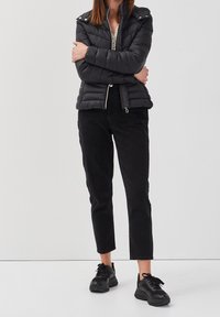 Doudoune noire avec fermeture éclair et accents de boutons-pression, associée à un pantalon en corduroy noir et des baskets noires. Designtexturé et moderne.