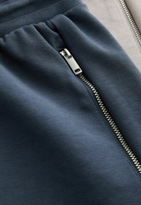 Gros plan sur une fermeture éclair en métal argenté partiellement fermée sur un tissu bleu foncé, mettant en valeur la texture et les détails de la couture.