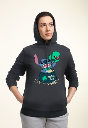 Osoba ubrana w czarną bluzę z kapturem z postacią z kreskówki, trzymająca zielony kapelusz skrzata i garnek oznaczony napisem "Lucky Me" wypełniony złotymi monetami.