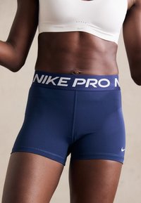 Marineblå Nike Pro shorts med en bred talje, der har hvid "NIKE PRO" tekst. Glat stof, tætsiddende design og ingen synlige mønstre.