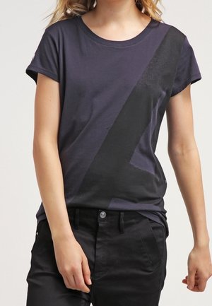 Femme portant un t-shirt gris foncé avec une large bande diagonale noire, assorti à un pantalon noir, debout avec une posture détendue.