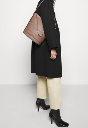 Sac seau en cuir marron avec accent chaîne, texture lisse et forme structurée, porté avec un manteau noir et un pantalon de couleur crème.