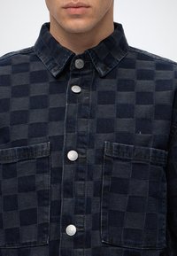 Homme portant une chemise à carreaux bleu foncé boutonnée avec deux poches poitrine, boutons argentés et col, visible du cou jusqu'à la mi-torse.
