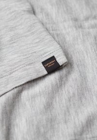 Šedé tričko z jemné látky, s hladkou texturou, se černou nášivkou s nápisem "SUPERDRY" zlatým písmem na rukávu.