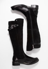 Bottes noires montantes jusqu'au genou, combinant cuir lisse et suède. Fermeture éclair sur le côté, détail avec sangle en cuir, et petit talon.