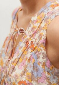 Blouse sans manches à motif floral dans des couleurs pastel, avec un tissu doux, un détail de nœud au niveau du col et de légères plis.