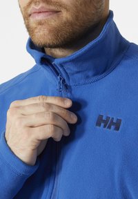 Blauwe fleece jacket met een hoge kraag, ritssluiting aan de voorkant en een geborduurd logo in donkerblauw op de borst. Zachte textuur is zichtbaar.