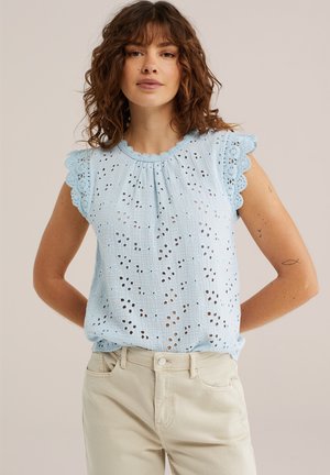 Žena s kučeravými hnedými vlasmi nosí svetlomodré tielko bez rukávov s dierkami a béžové nohavice, stojí pred jednofarebným béžovým pozadím.