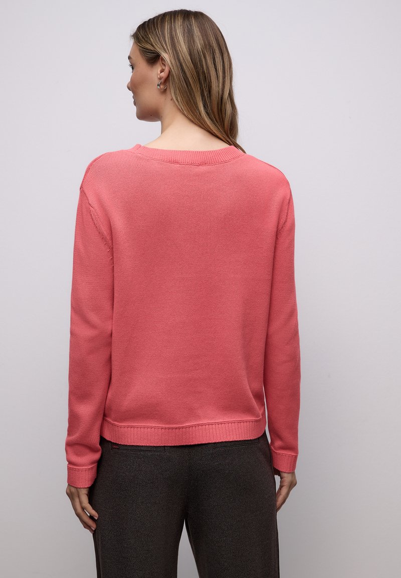 Pull en maille rose à manches longues, col et ourlet côtelés, de coupe décontractée. Porté avec un pantalon gris foncé, vu de dos.