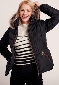 Schwarze Pufferjacke mit einer mit Fell gefütterten Kapuze, Reißverschluss vorne und braunen Akzenten. Getragen über einem gestreiften Strickpullover mit gerippten Manschetten.